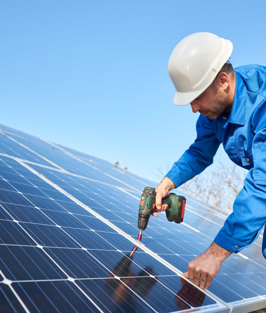 Man worker in blue suit and protective helmet installing solar photovoltaic panel system using screwdriver. Professional electrician mounting blue solar module. Alternative energy ecological concept.